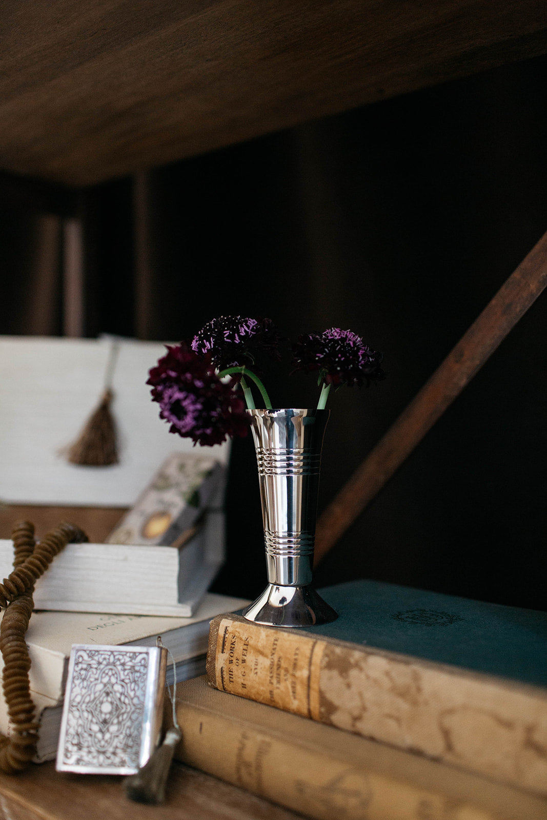 A silver metal vase with a flared top holding two dark purple flowers, styled on a wooden shelf with vintage books and a small silver card.
